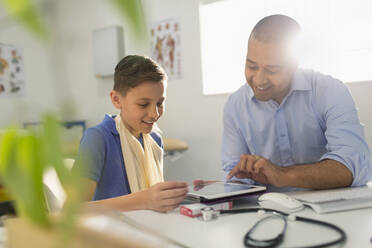 Ein männlicher Kinderarzt zeigt einem jungen Patienten in einer Arztpraxis ein digitales Tablet - CAIF25405