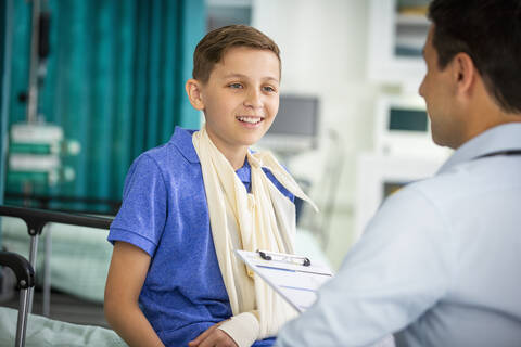 Männlicher Kinderarzt im Gespräch mit einem jungen Patienten mit Arm in einer Schlinge in einer Klinik, lizenzfreies Stockfoto