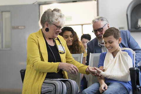 Eine Kinderärztin untersucht die Hand eines Jungen mit einem Arm in einer Schlinge im Foyer einer Klinik, lizenzfreies Stockfoto