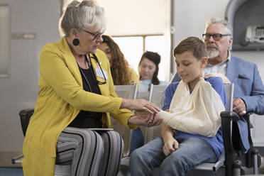 Female doctor examining hand of boy patient with arm in sling in clinic lobby - CAIF25472