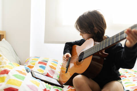 Boy sitting on bed using digital tablet for playing song on guitar stock photo