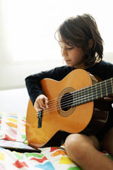 Junge, der auf dem Bett sitzt und ein digitales Tablet benutzt, um ein Lied auf der Gitarre zu spielen - VABF02689