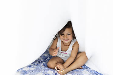 Porträt eines kleinen Mädchens, das auf dem Bett sitzt und sich unter einer Decke versteckt - VABF02709