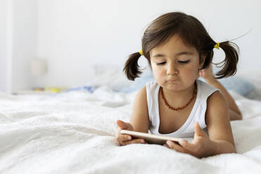 Portrait of little girl lying on bed in underwear looking at smartphone - VABF02712