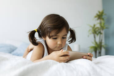 Porträt eines kleinen Mädchens, das in Unterwäsche auf dem Bett liegt und auf sein Smartphone schaut - VABF02716