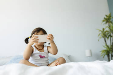Kleines Mädchen sitzt in Unterwäsche auf dem Bett und benutzt ein Smartphone - VABF02718
