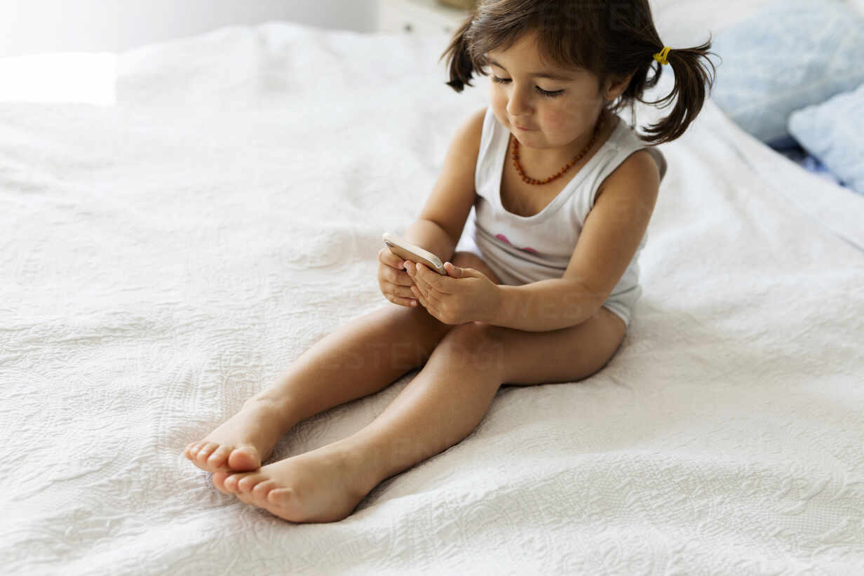 Little girl in underwear sitting on bed looking at smartphone stock photo
