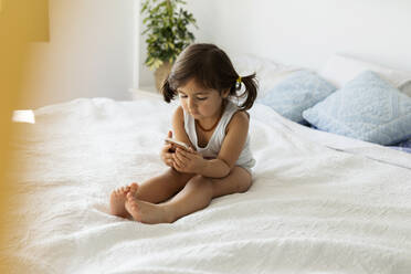 Little girl in underwear sitting on bed looking at smartphone - VABF02723