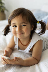 Portrait of smiling little girl with smartphone lying on bed - VABF02725