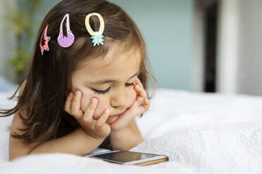 Porträt eines kleinen Mädchens, das auf dem Bett liegt und auf sein Smartphone schaut - VABF02728