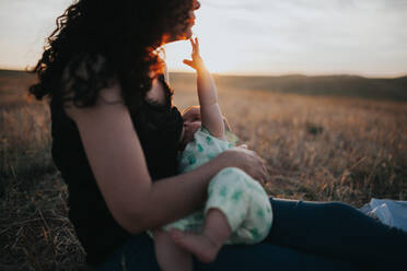 Seitenansicht der Mutter, die ihr Baby stillt, während sie auf einem Feld gegen den Himmel bei Sonnenuntergang sitzt - EYF02179