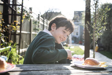 Ssmiling boy wearing green jumper sitting at a table in a garden with a snack. - CUF54950