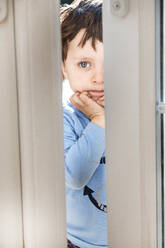 Porträt eines Jungen in blauem Hemd, der durch eine leicht geöffnete Tür blickt. - CUF54953