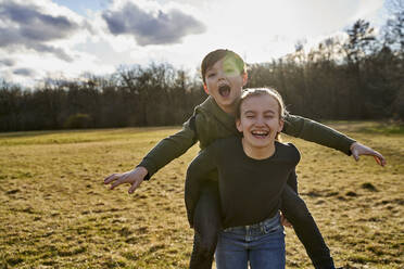 Happy girl carrying brother piggyback on a meadow - AUF00278