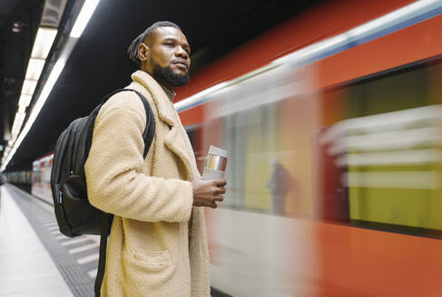 Stylish man with reusable cup and headphones in a metro station - AHSF02116
