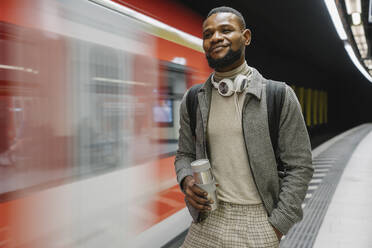 Stilvoller Mann mit wiederverwendbarem Becher und Kopfhörern in einer U-Bahn-Station - AHSF02119