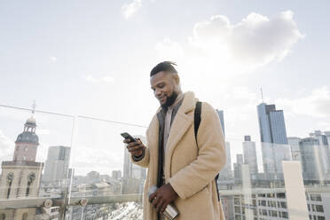 Stylischer Mann mit Smartphone auf einer Aussichtsterrasse mit Blick auf ein Hochhaus, Frankfurt, Deutschland - AHSF02127