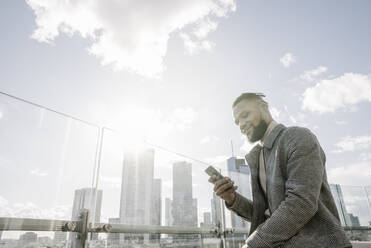 Stylischer Mann mit Smartphone auf einer Aussichtsterrasse mit Blick auf ein Hochhaus, Frankfurt, Deutschland - AHSF02132