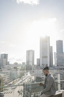 Stylischer Mann mit Smartphone auf einer Aussichtsterrasse mit Blick auf ein Hochhaus, Frankfurt, Deutschland - AHSF02133