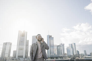 Lächelnder Mann am Telefon auf einer Aussichtsterrasse mit Blick auf ein Hochhaus, Frankfurt, Deutschland - AHSF02142