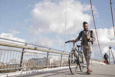Stilvoller Mann mit einem Fahrrad, der sein Smartphone benutzt, während er auf einer Brücke geht - AHSF02152