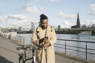 Stylischer Mann mit Fahrrad und Smartphone am Flussufer, Frankfurt, Deutschland - AHSF02164