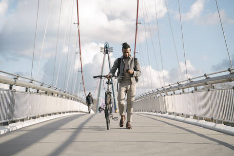 Stylish man with a bicycle using smartphone while walking on a bridge stock photo