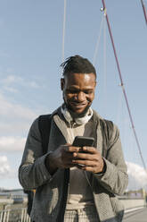 Porträt eines lächelnden Mannes auf einer Brücke mit Kopfhörern und Smartphone - AHSF02168