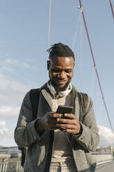 Portrait of smiling man on a bridge with headphones using smartphone - AHSF02168