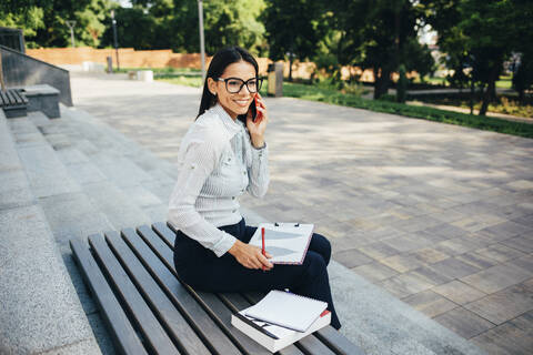 Lächelnde Geschäftsfrau, die anruft und auf einer Bank in einem Park sitzt, lizenzfreies Stockfoto