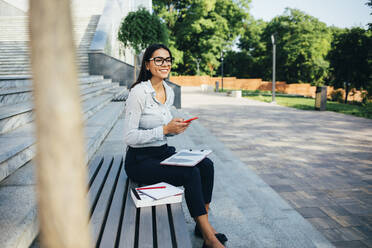 Businesswoman using smartphone and sitting on a bench in a park - OYF00117