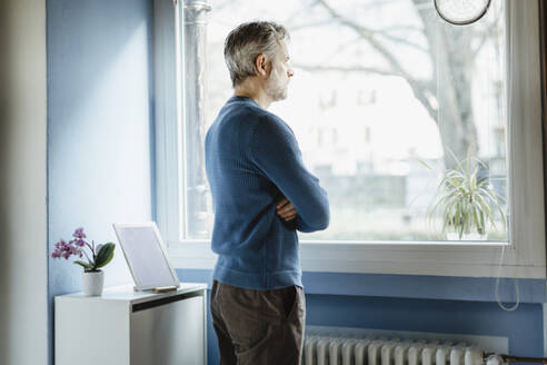 Pensive man standing in living room looking out of window - MCVF00248