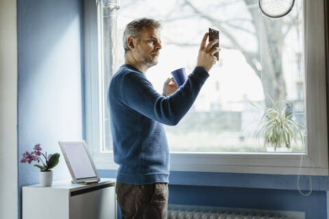 Älterer Mann mit Kaffeetasse im Wohnzimmer stehend, der ein Selfie mit seinem Smartphone macht, lizenzfreies Stockfoto