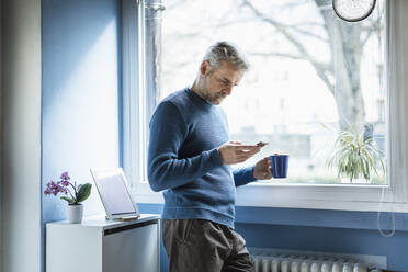 Älterer Mann mit Kaffeetasse im Wohnzimmer stehend, der auf sein Smartphone schaut - MCVF00250
