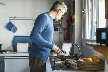 Älterer Mann bei der Zubereitung von Artischocken in seiner Küche mit einem digitalen Tablet - MCVF00254