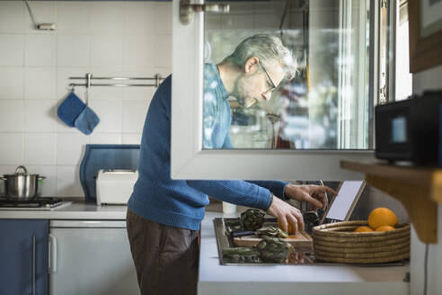 Mature man preparing artichoke in his kitchen using digital tablet - MCVF00255