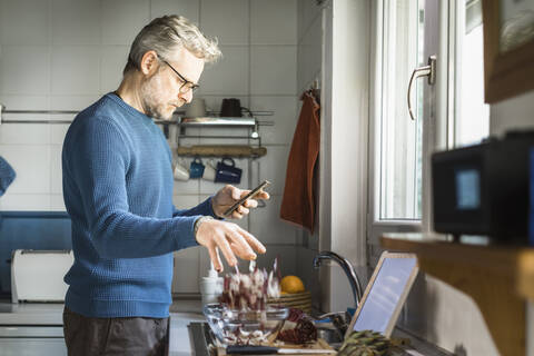 Älterer Mann steht in seiner Küche und schaut auf sein Smartphone, während er einen Salat zubereitet, lizenzfreies Stockfoto