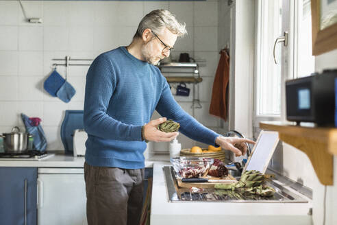 Mature man preparing salad in his kitchen using digital tablet - MCVF00259