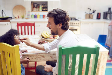 Father feeding daughter grapes at kitchen table - CAIF25505