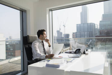 Nachdenklicher Geschäftsmann mit Papierkram in einem sonnigen, modernen, städtischen Büro - CAIF25590