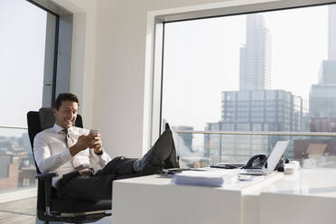 Smiling businessman using smart phone with feet up on desk in modern, sunny, urban office - CAIF25594