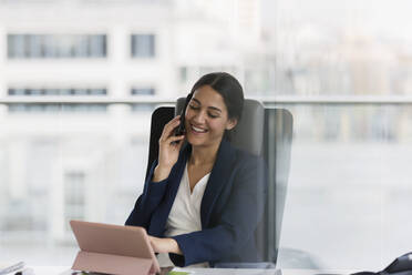 Lächelnde Geschäftsfrau, die im Büro mit einem Smartphone telefoniert - CAIF25624