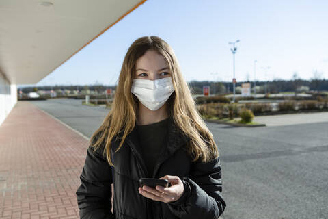 Porträt eines Mädchens mit Maske auf einer leeren Straße, das ein Smartphone benutzt, lizenzfreies Stockfoto