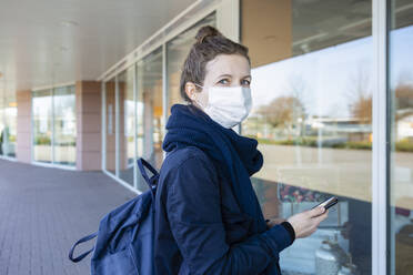 Frau mit Maske, die vor einem Schaufenster eines geschlossenen Einkaufszentrums steht und Smarthone benutzt - OJF00375