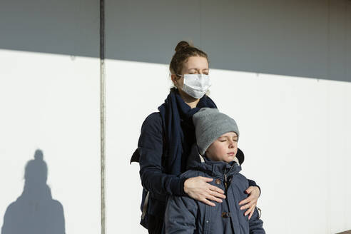 Woman wearing mask standing with son in front of a white wall, eyes closed - OJF00376