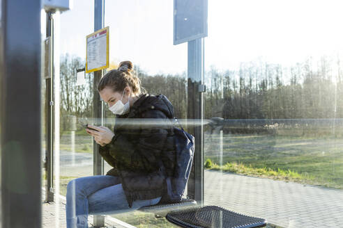 Girl with mask waiting at bus stop, using smartphone - OJF00379
