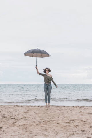 Junge Frau springt am Strand und hält einen Regenschirm, lizenzfreies Stockfoto