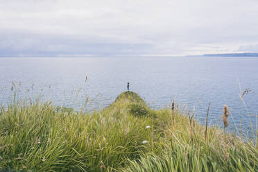 Junge Frau steht im Gras an der Küste und schaut auf das Meer - FVSF00021