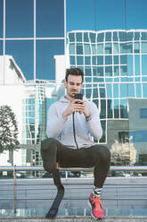 Disabled athlete with leg prosthesis having a break from exercising in the city using cell phone - DAMF00300