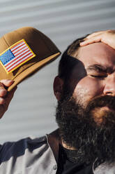 Bärtiger Mann mit Baseballmütze mit amerikanischer Flagge wischt sich den Schweiß vom Kopf - JCMF00555